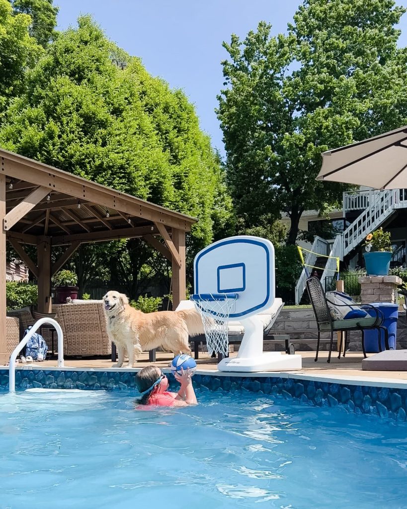 Young girl in a swimming pool, poised to throw a pool basketball into the nearby hoop, capturing the spirit of summertime fun.